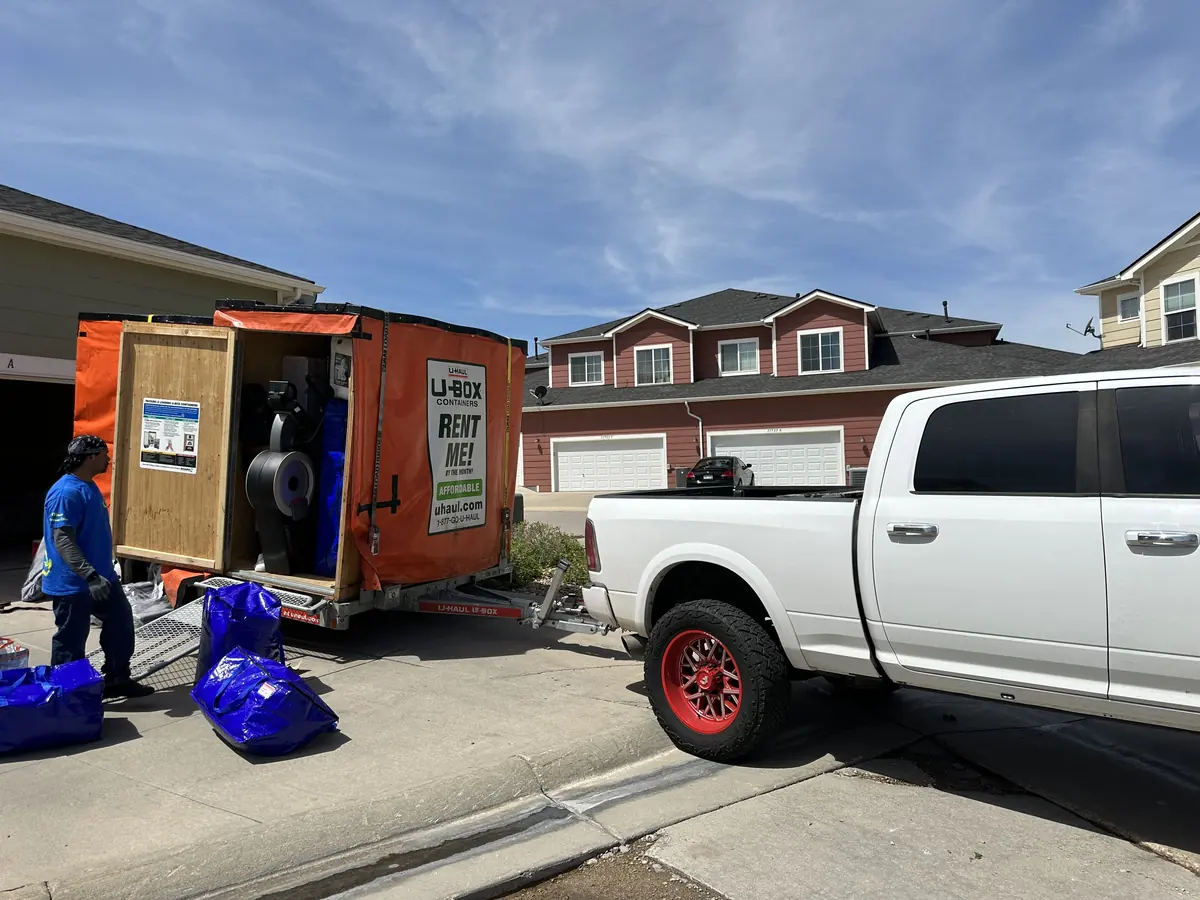 Mover beside an open box truck and a white pickup at a residential driveway